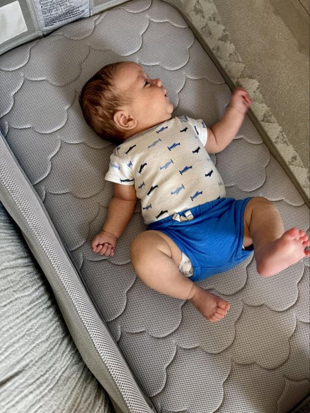 Infant lying on a gray quilted mattress, wearing a patterned white shirt and blue shorts.