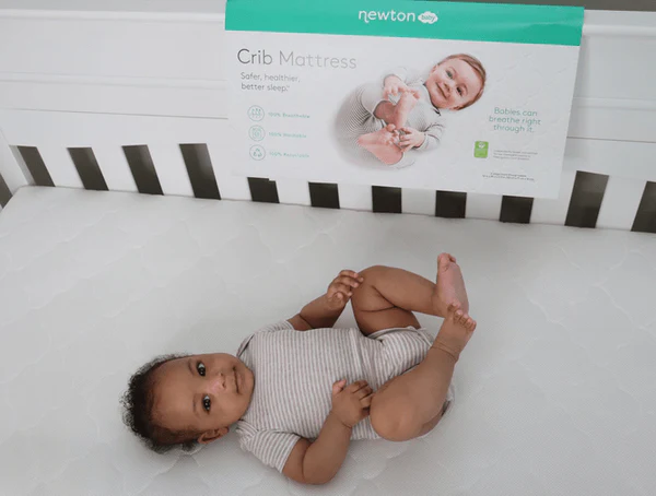 Baby lying on a white crib mattress with a Newton Crib Mattress box behind them.