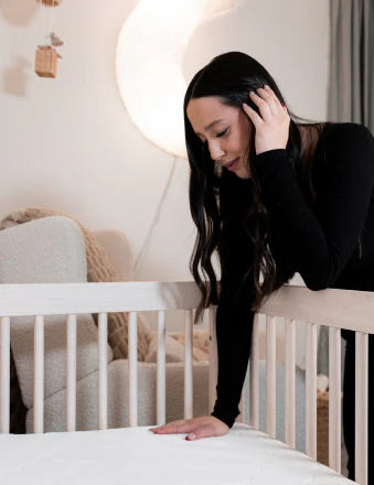 Woman leaning over a white crib with one hand on the mattress in a softly lit nursery.