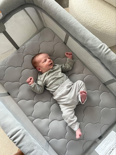 Newborn lying on a gray quilted pad inside a mesh-sided bassinet