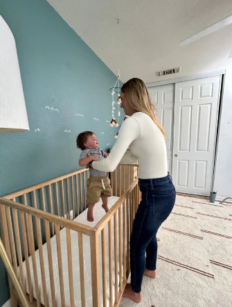 Adult woman steadying a young child standing in a wooden crib in a nursery.