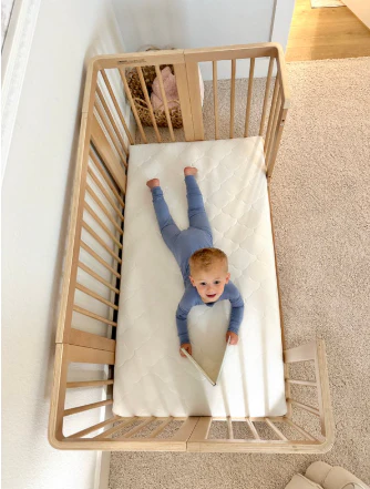 Baby in blue pajamas lying on a white crib mattress inside a light wood crib.