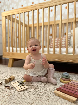 Baby sitting on a carpet in front of a wooden crib with toys nearby
