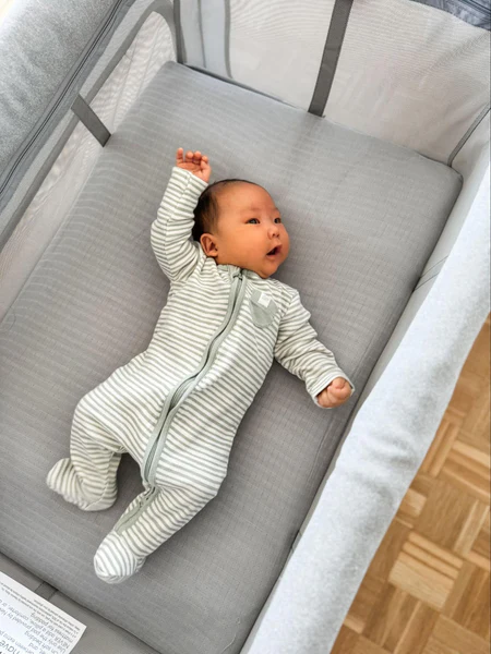 Infant in a striped footed sleeper lying on a gray mattress with one arm raised