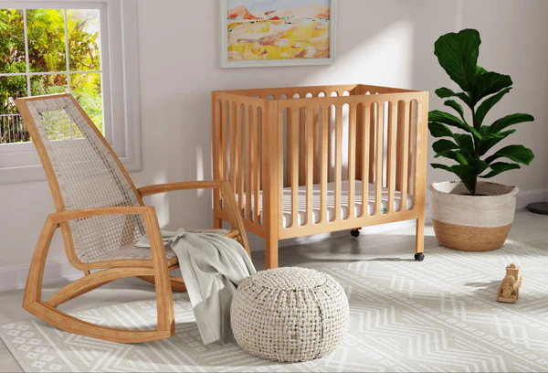 Wooden crib beside a rocking chair, pouf, potted plant, and window in a bright nursery