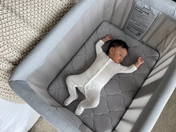 Infant in white footed pajamas lying on a gray quilted mattress inside a light gray bassinet.