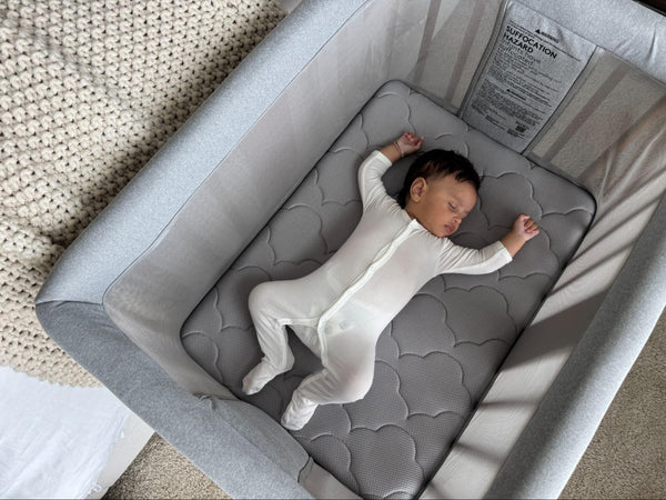 Infant sleeping on their back in a white sleeper on a gray mattress inside a light gray bassinet.