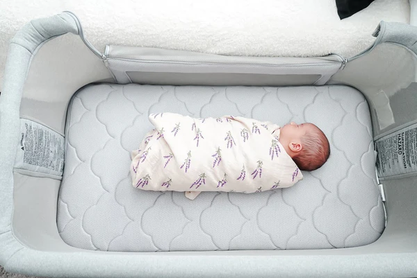 Swaddled baby lying on a quilted mattress inside a light gray bassinet