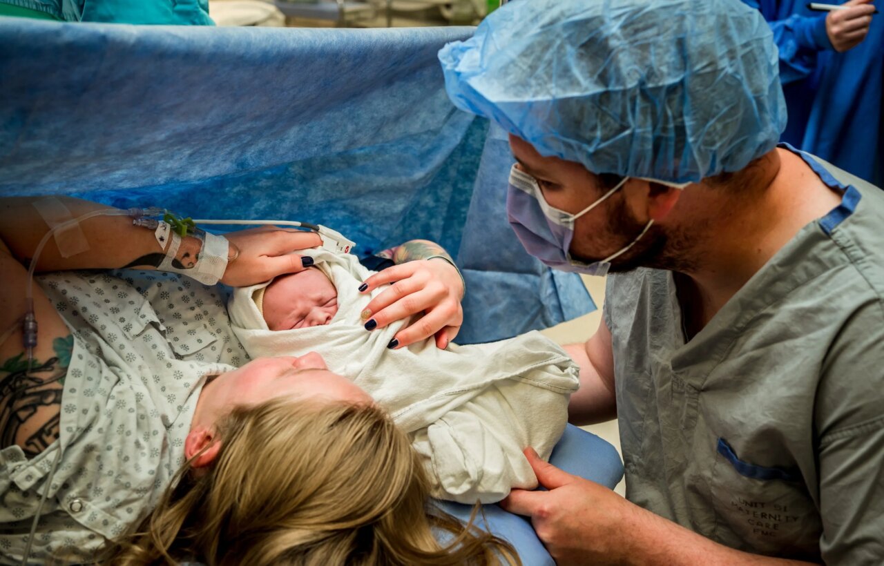 Newborn wrapped in a blanket held beside parents and medical staff in an operating room