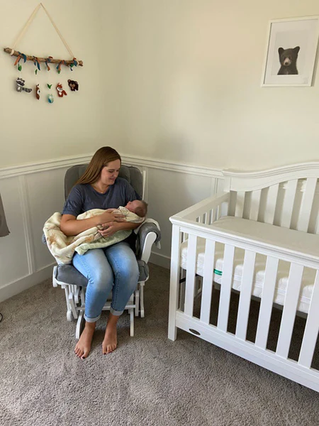 Adult seated in a nursery holding a baby beside a white crib.