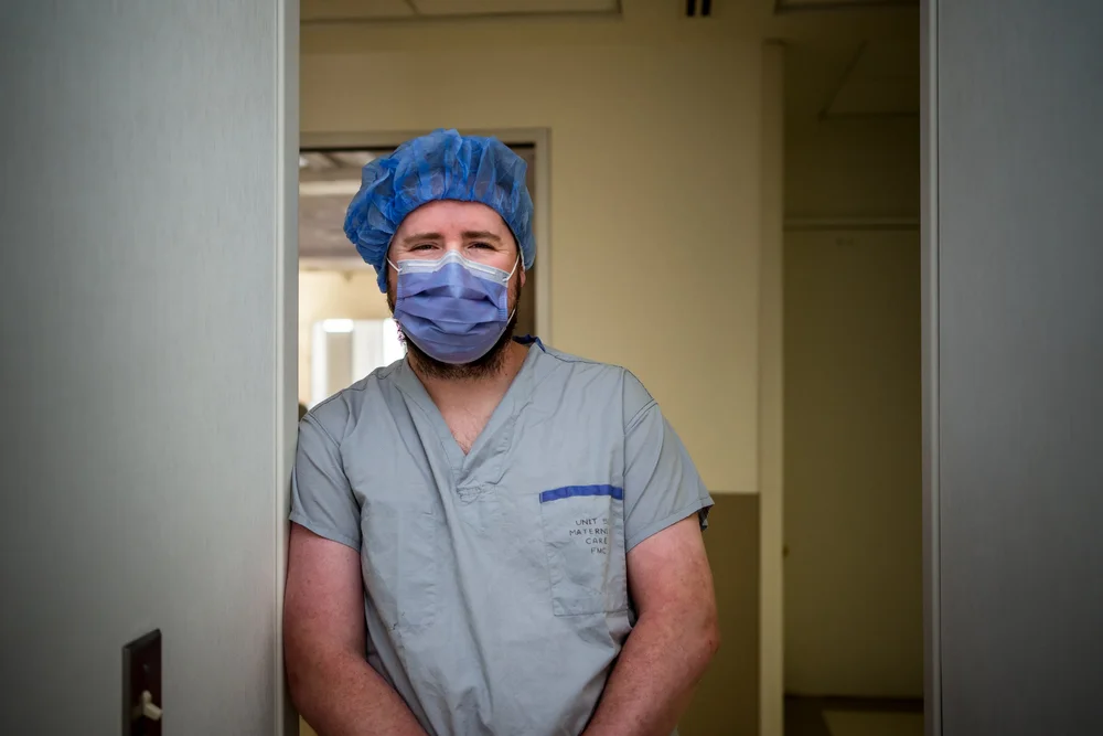 Masked person in scrubs and blue hair cover standing in a doorway