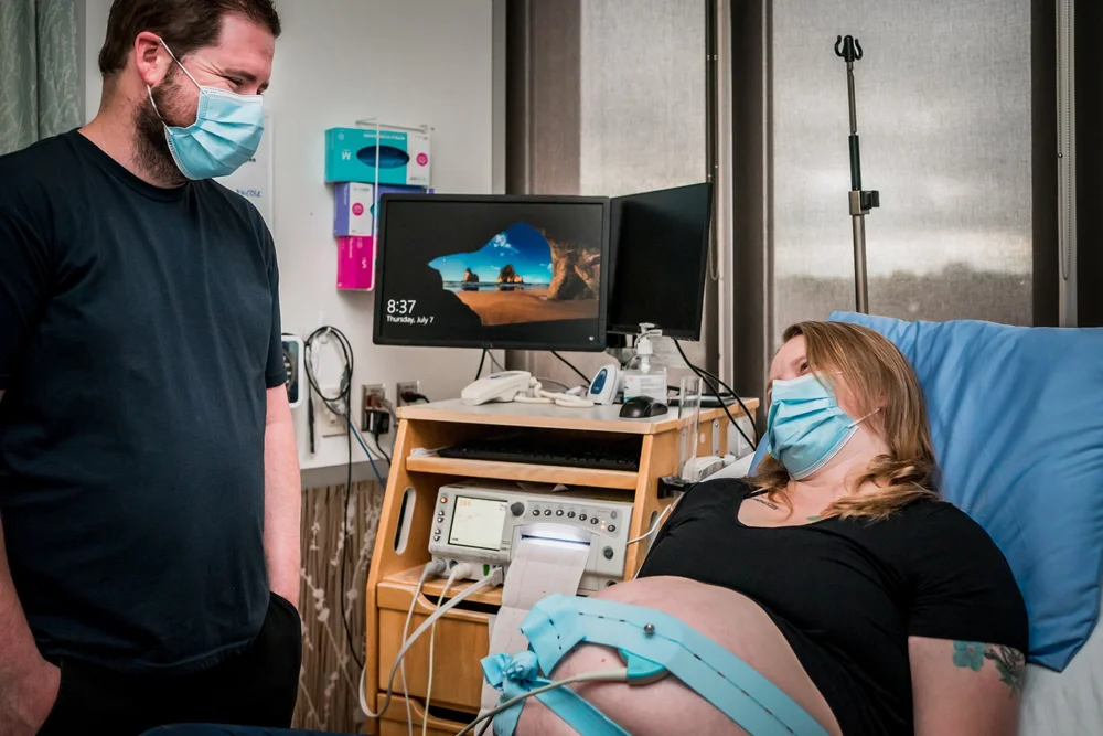 Pregnant patient wearing a mask reclines in a hospital bed beside a masked support person.