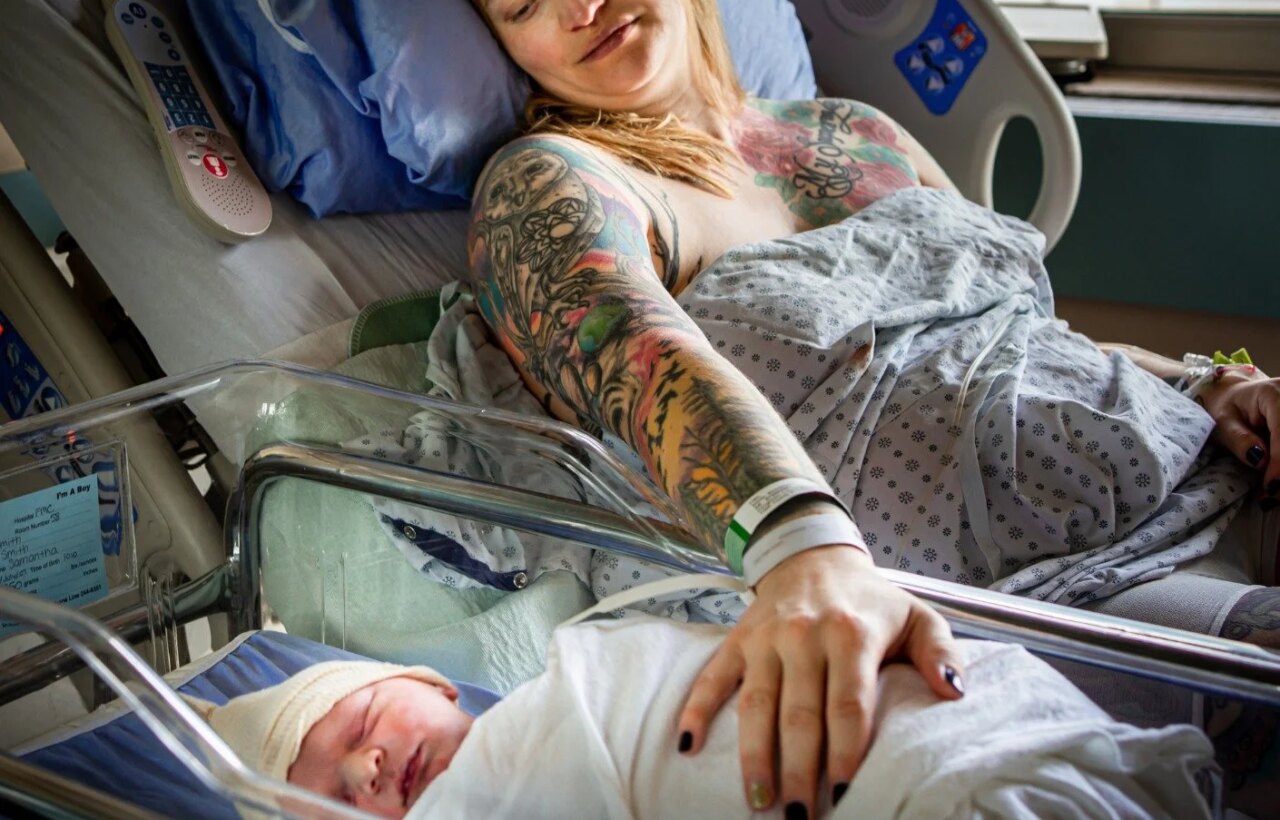 Woman in a hospital bed rests her hand on a swaddled newborn in a bassinet.