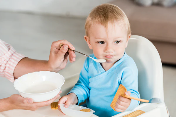 Young child in a high chair being fed with a spoon while holding a cracker and spoon.