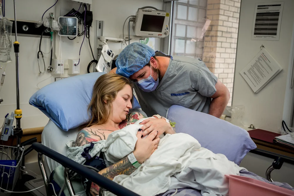 New mother cradles a swaddled baby in a hospital bed while a masked person leans close beside her.