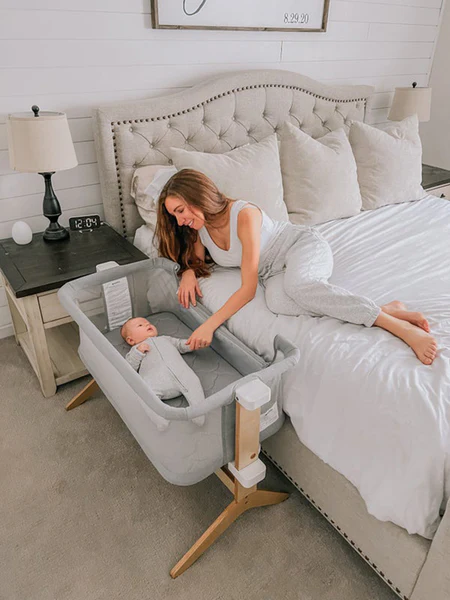 Woman on a bed reaching toward an infant lying in a bedside bassinet beside a nightstand.