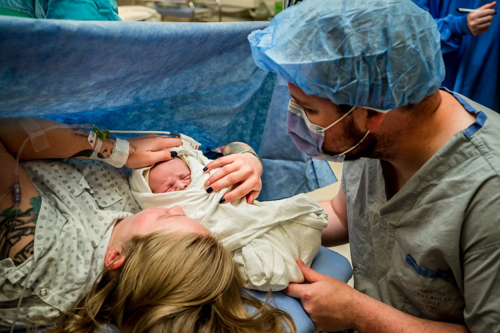 Newborn wrapped in a blanket held near a parent in surgical clothing in an operating room