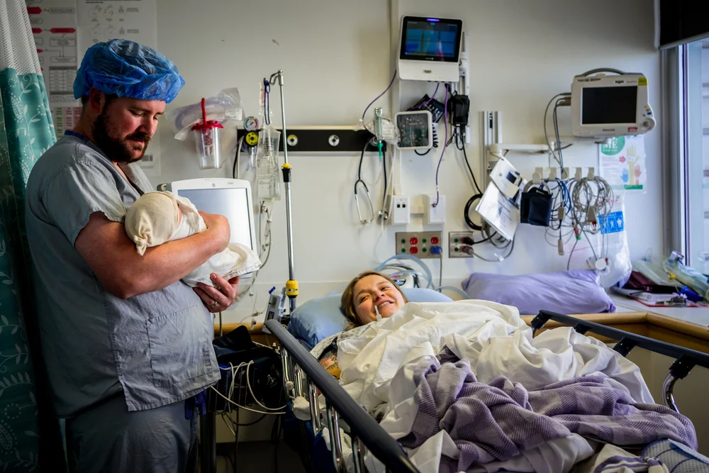 Person holding a swaddled newborn beside a smiling patient in a hospital bed