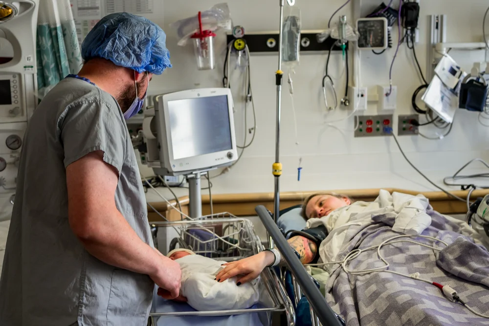 Scrub-clad person beside a wrapped newborn while a patient rests in a hospital bed.