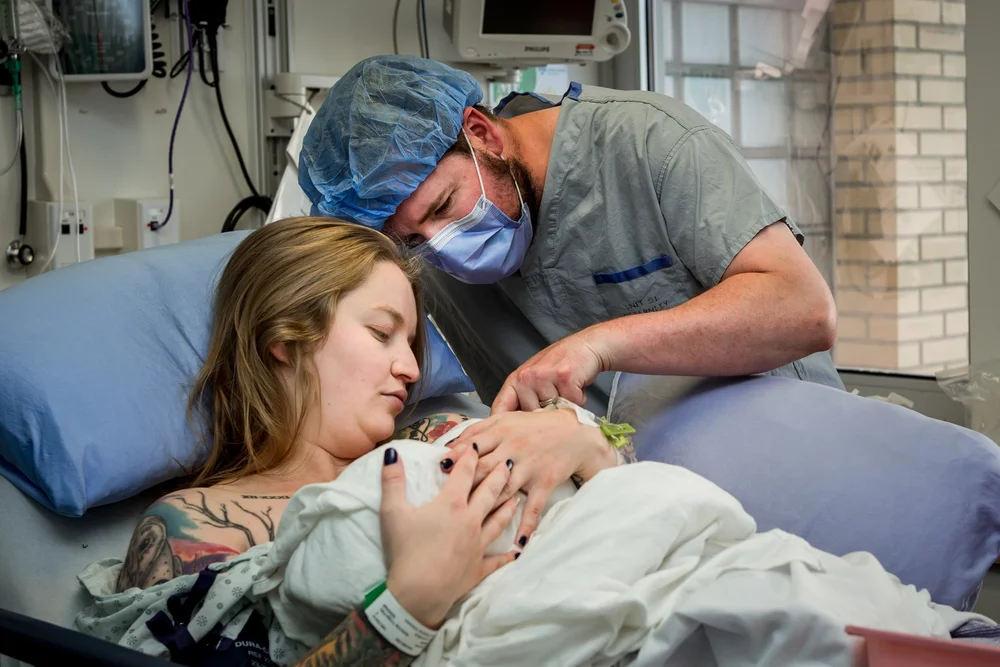 Person in a hospital bed holding a wrapped baby while a masked person in scrubs leans close beside them.
