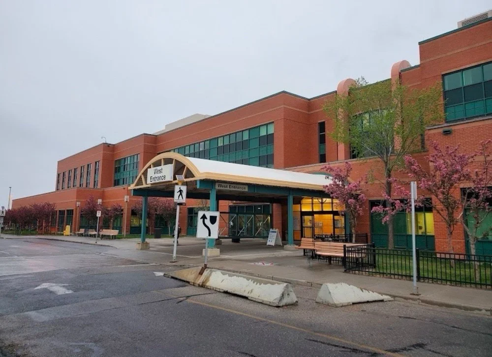 Brick hospital entrance with covered walkway, benches, trees, and signs reading West Entrance.