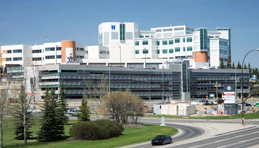 Large white hospital complex with glass parking structure, roadway, trees, and blue H signs under a clear sky