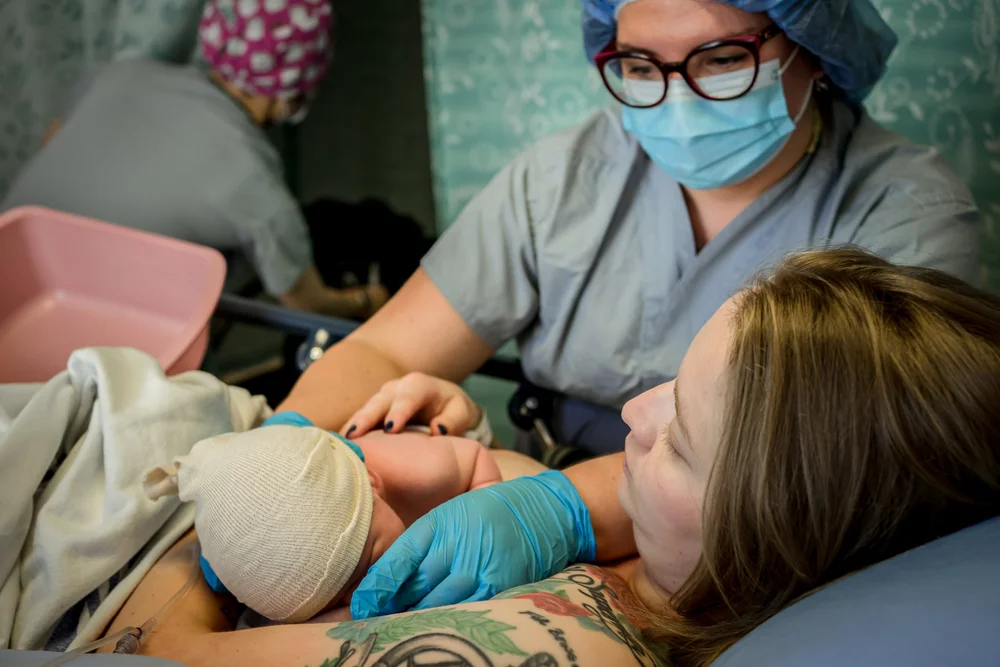 Newborn in a knit cap lies on an adult's chest while a masked medical worker and gloved hands provide care.