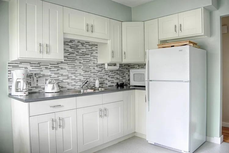 Small white kitchen with cabinets, sink, mosaic backsplash, refrigerator, microwave, coffee maker, and kettle