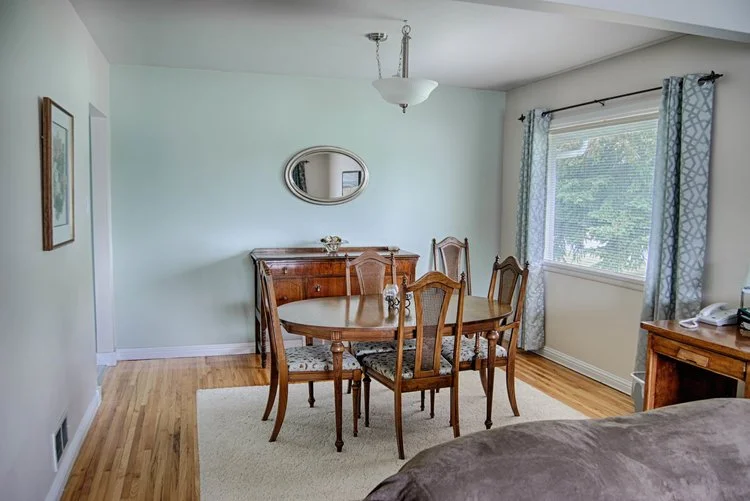 Small dining area with wooden table, chairs, sideboard, oval mirror, window, curtains, and hardwood floor.