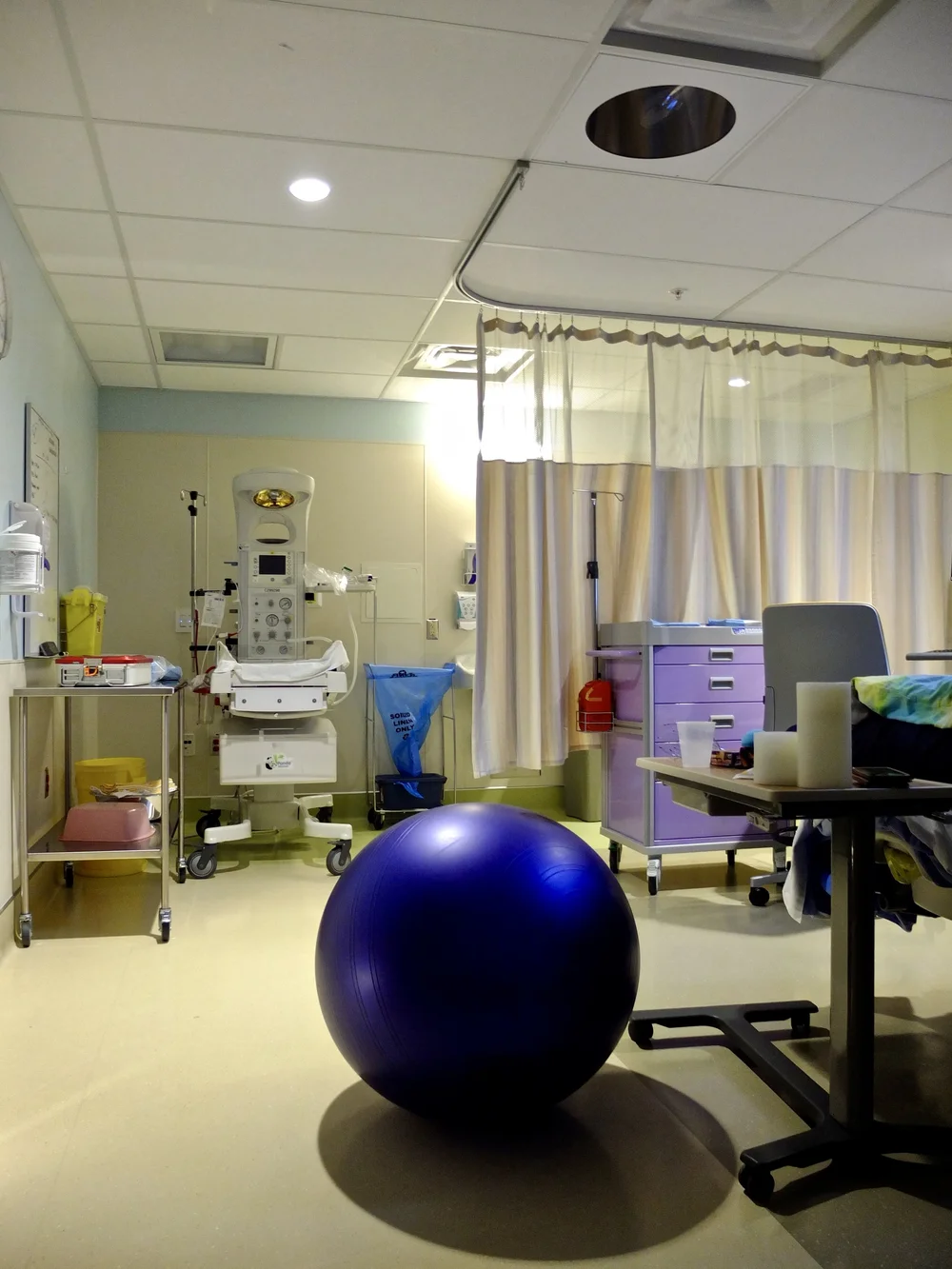 Hospital birth room with a blue exercise ball, curtains, infant warmer, carts, and medical equipment.