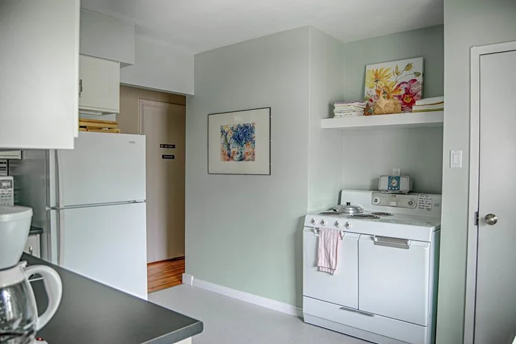 Small kitchen area with white refrigerator, stove, shelves, wall art, and a coffee maker in the foreground.