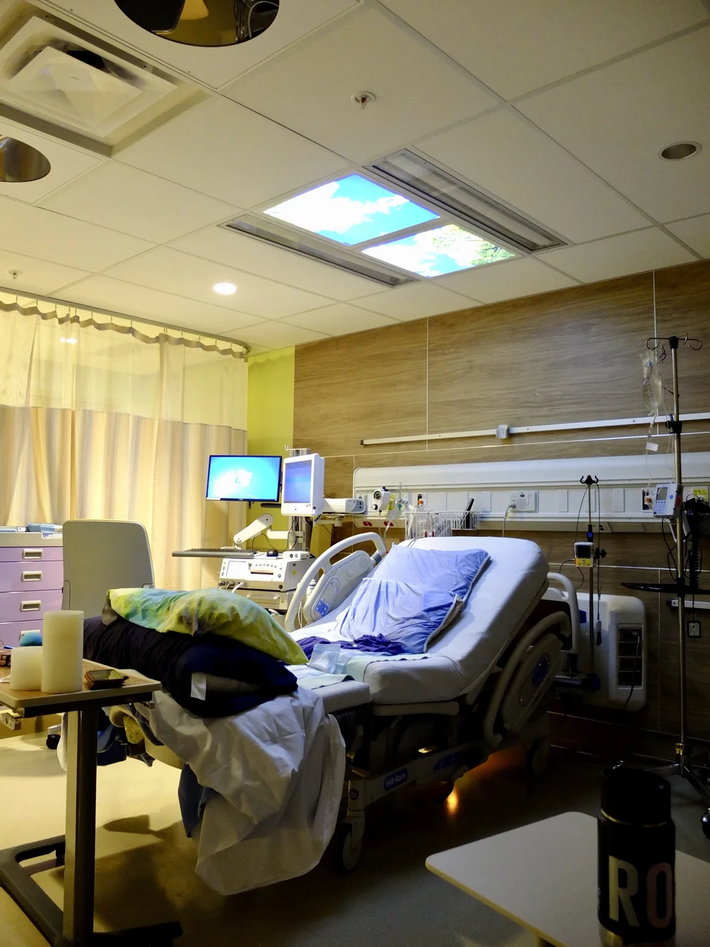 Hospital room with an adjustable bed, monitors, medical equipment, curtains, and a ceiling window.