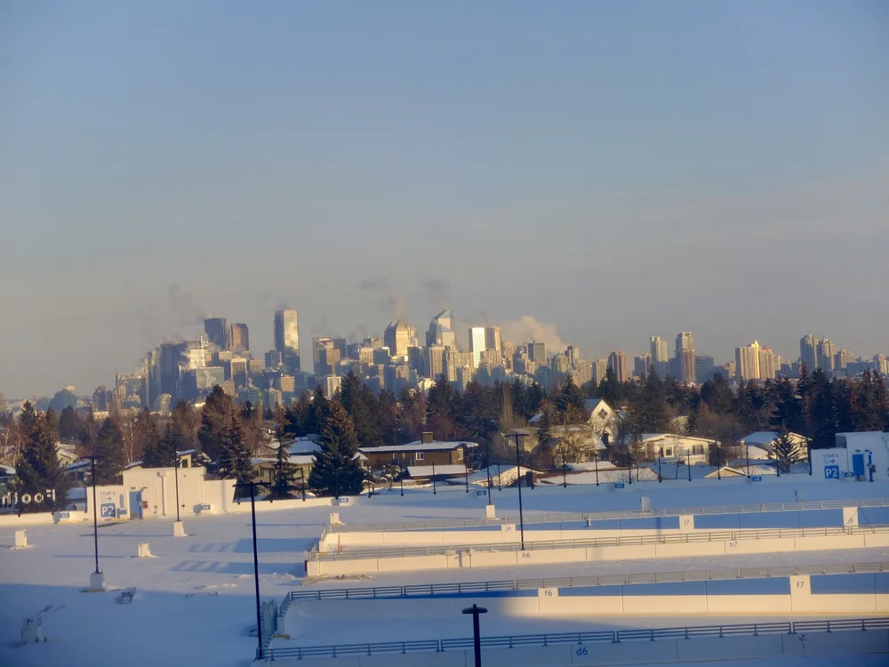 Snowy foreground with parking structures, trees, and a distant city skyline under a hazy blue sky.