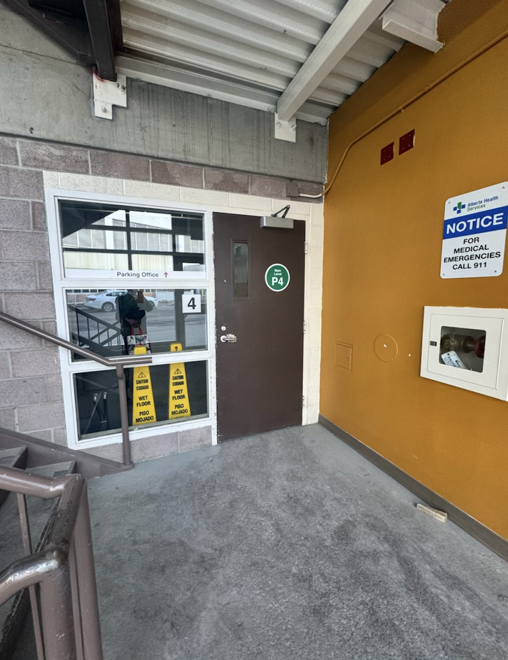 Covered parking level entrance with a P4 door, Parking Office window, stairs, and emergency notice sign.
