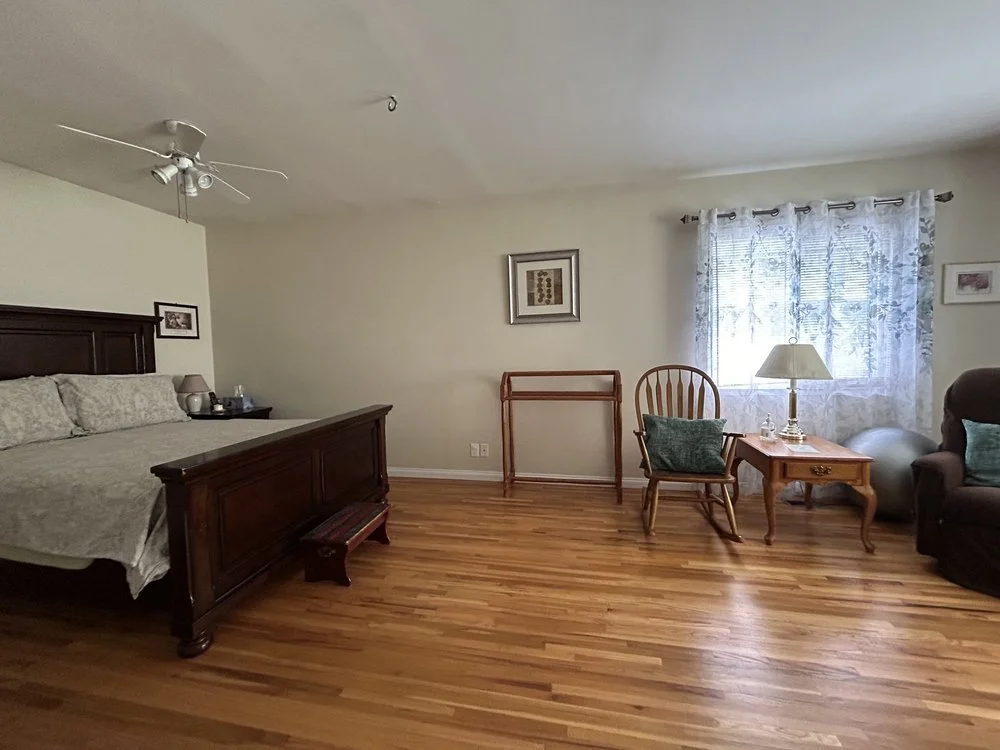 Spacious bedroom with wooden bed, hardwood floor, rocking chair, side table, lamp, window, and recliner