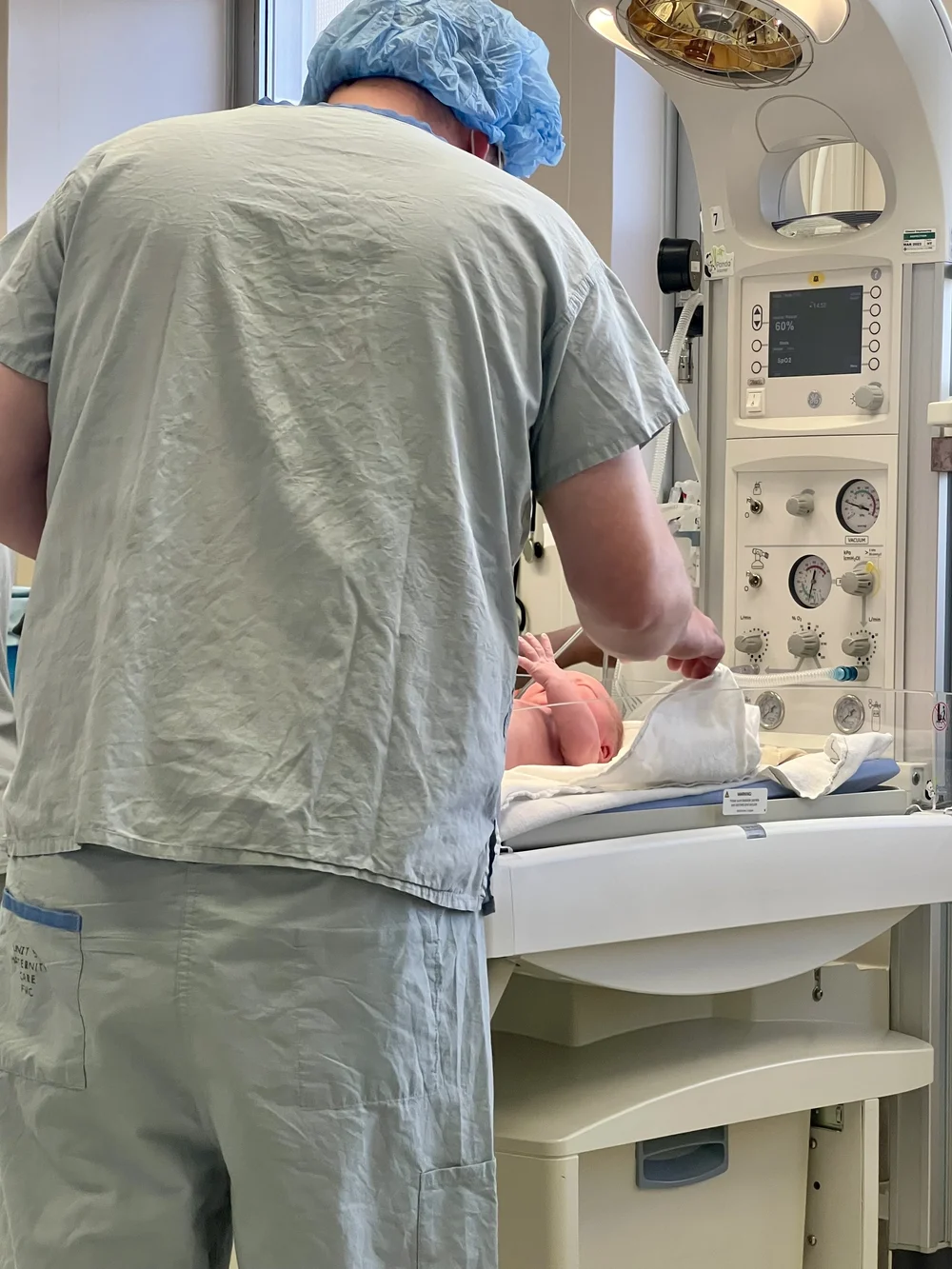 Person in surgical scrubs tending to an infant on a hospital warming table beside medical equipment