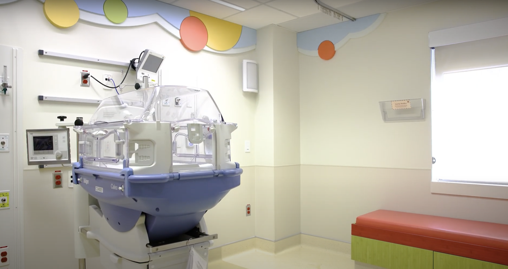 Hospital room with an infant incubator, wall equipment, colorful decor, and a bench by a window.