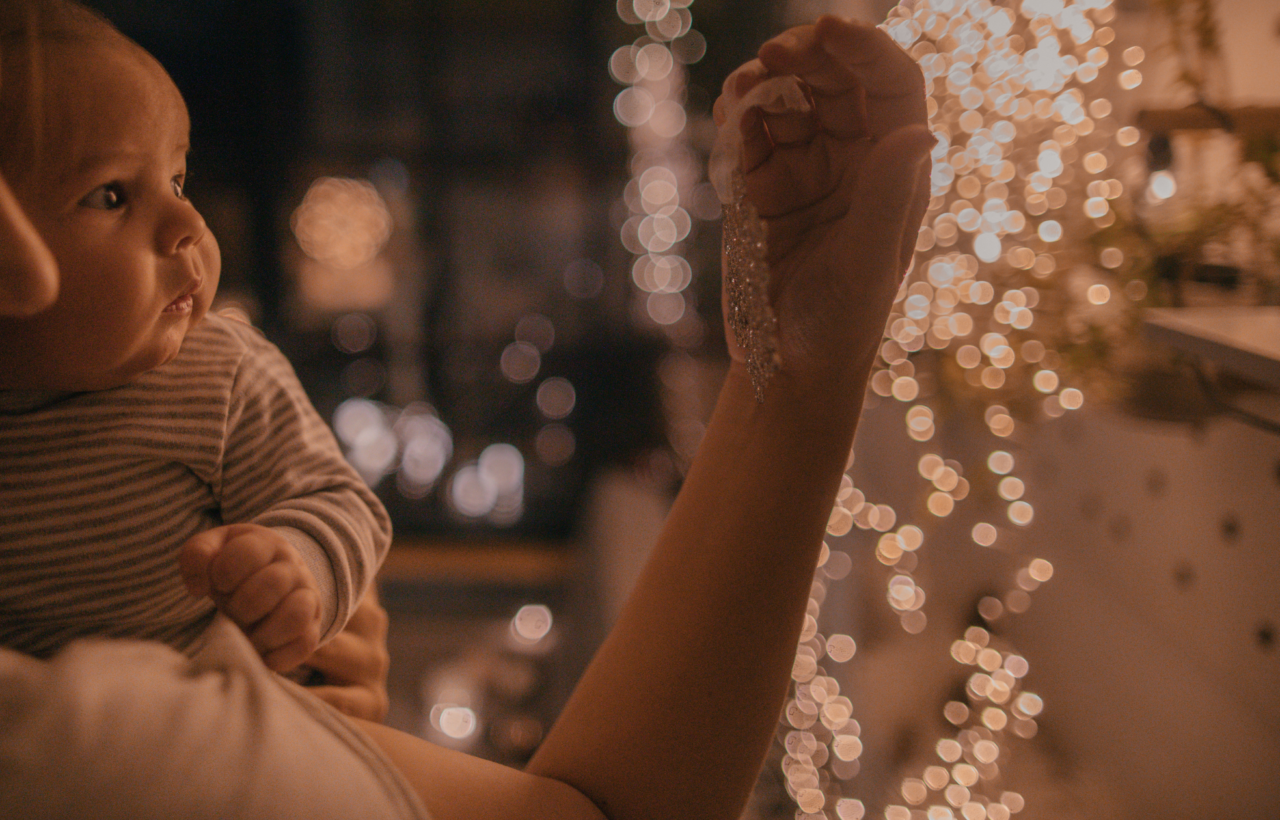 Baby and adult watching warm string lights beside a decorated tree indoors
