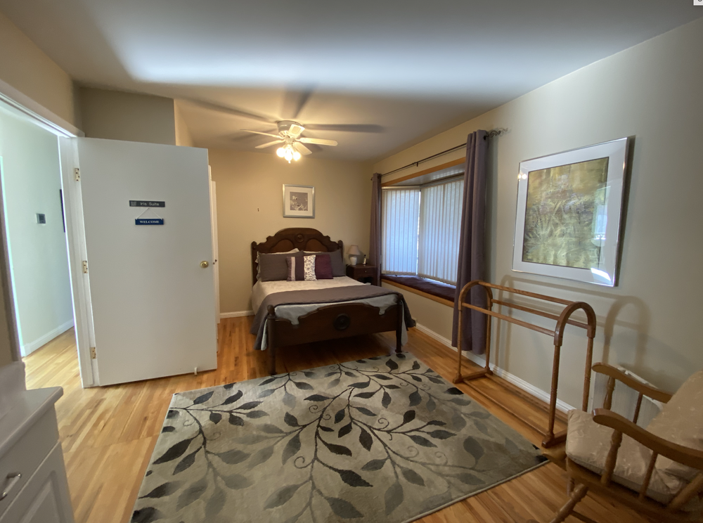 Bedroom with wooden bed, patterned rug, large window, rocking chair, and open door
