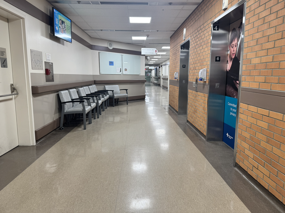 Hospital corridor with waiting chairs, elevators, brick wall, and a privacy notice panel.