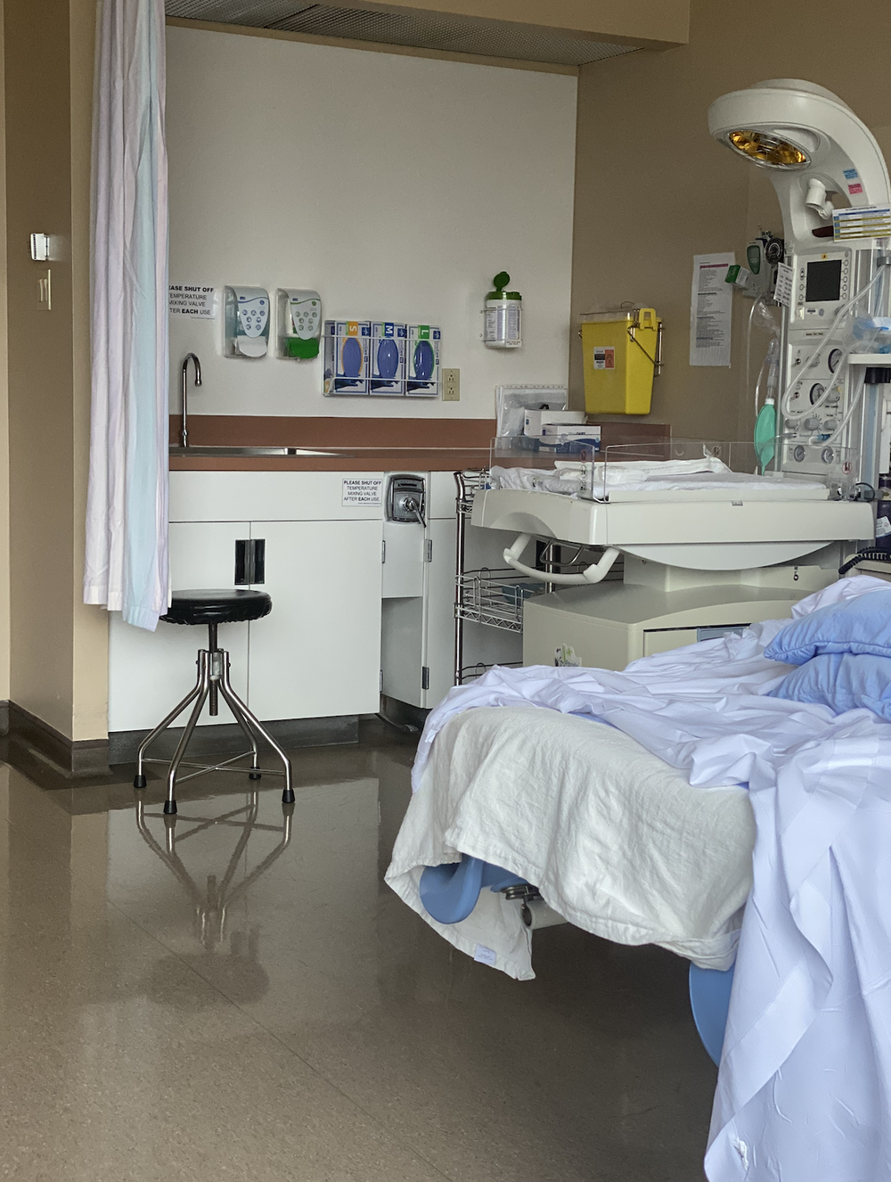 Hospital room with a bed, infant warmer, sink area, stool, curtain, and wall-mounted medical supplies.
