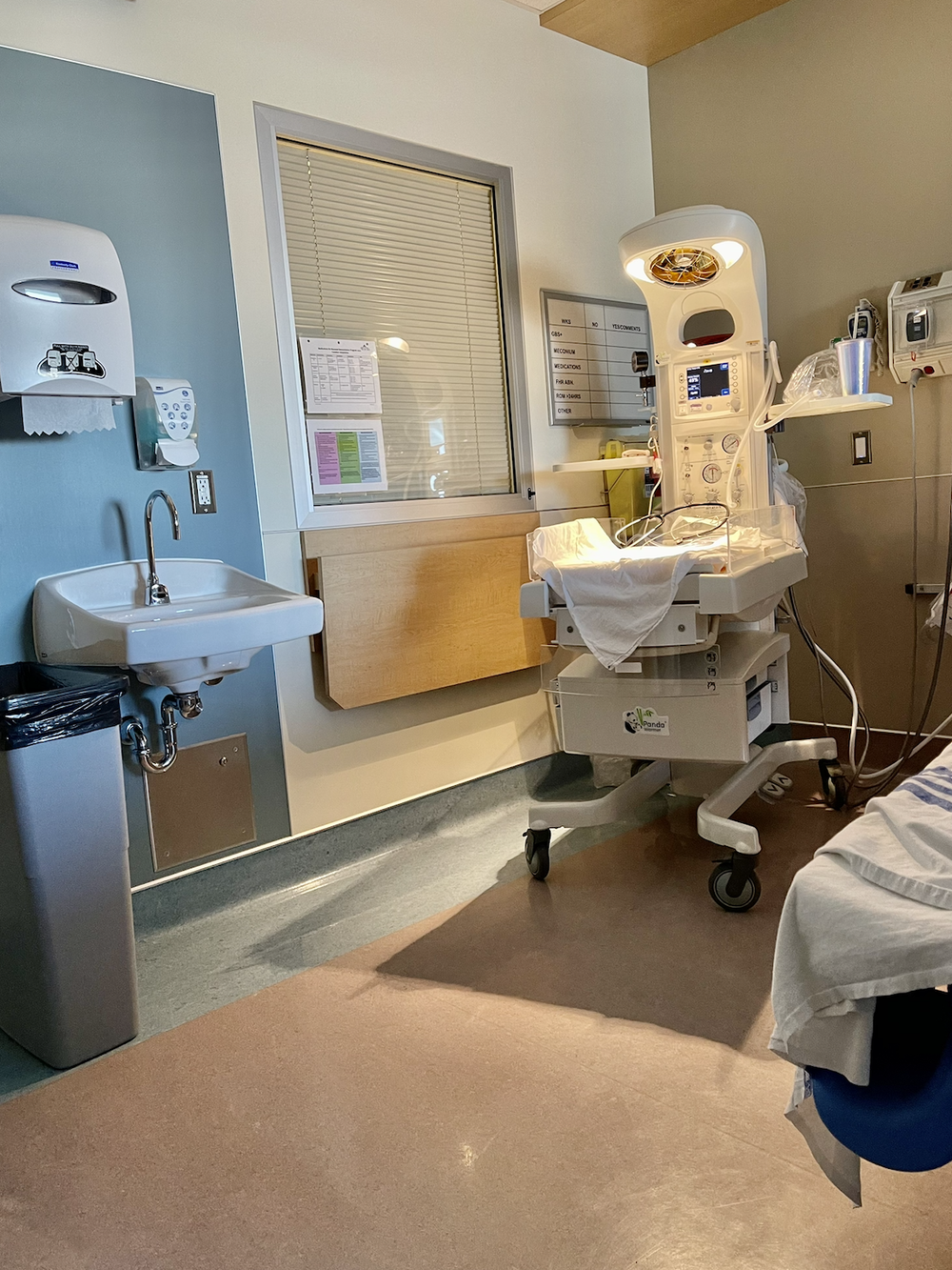 Hospital room with sink, window blinds, bed edge, and a neonatal warmer with lights and monitors.