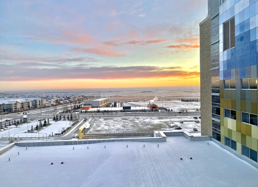 Snowy view from a building toward roads, low buildings, open fields, and a colorful sunset sky.