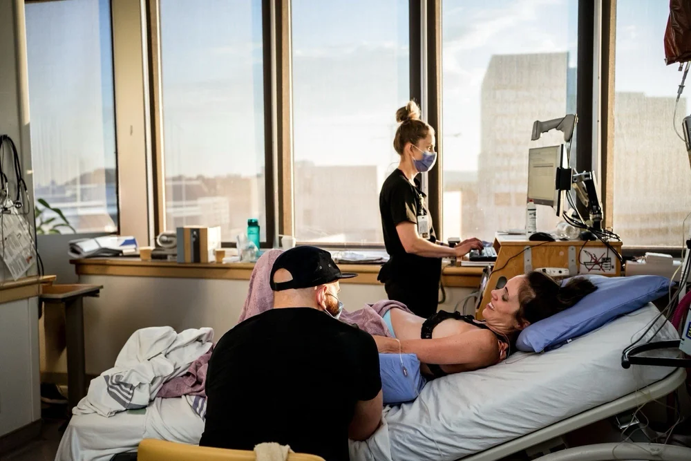 Patient in a hospital bed with a seated support person and a masked staff member at a computer nearby