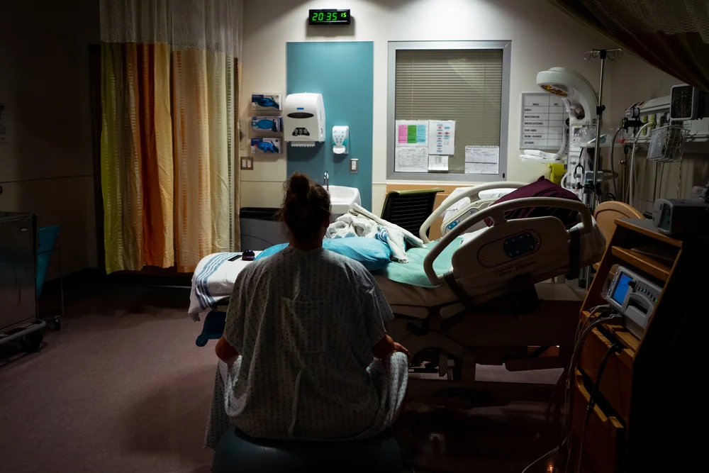Person in a hospital gown sits beside a hospital bed in a dimly lit room with medical equipment.
