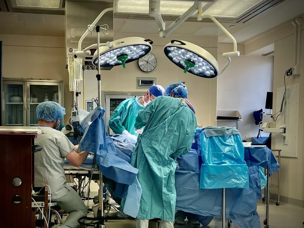 Medical team in surgical gowns gathered around a draped operating table under bright surgical lights.