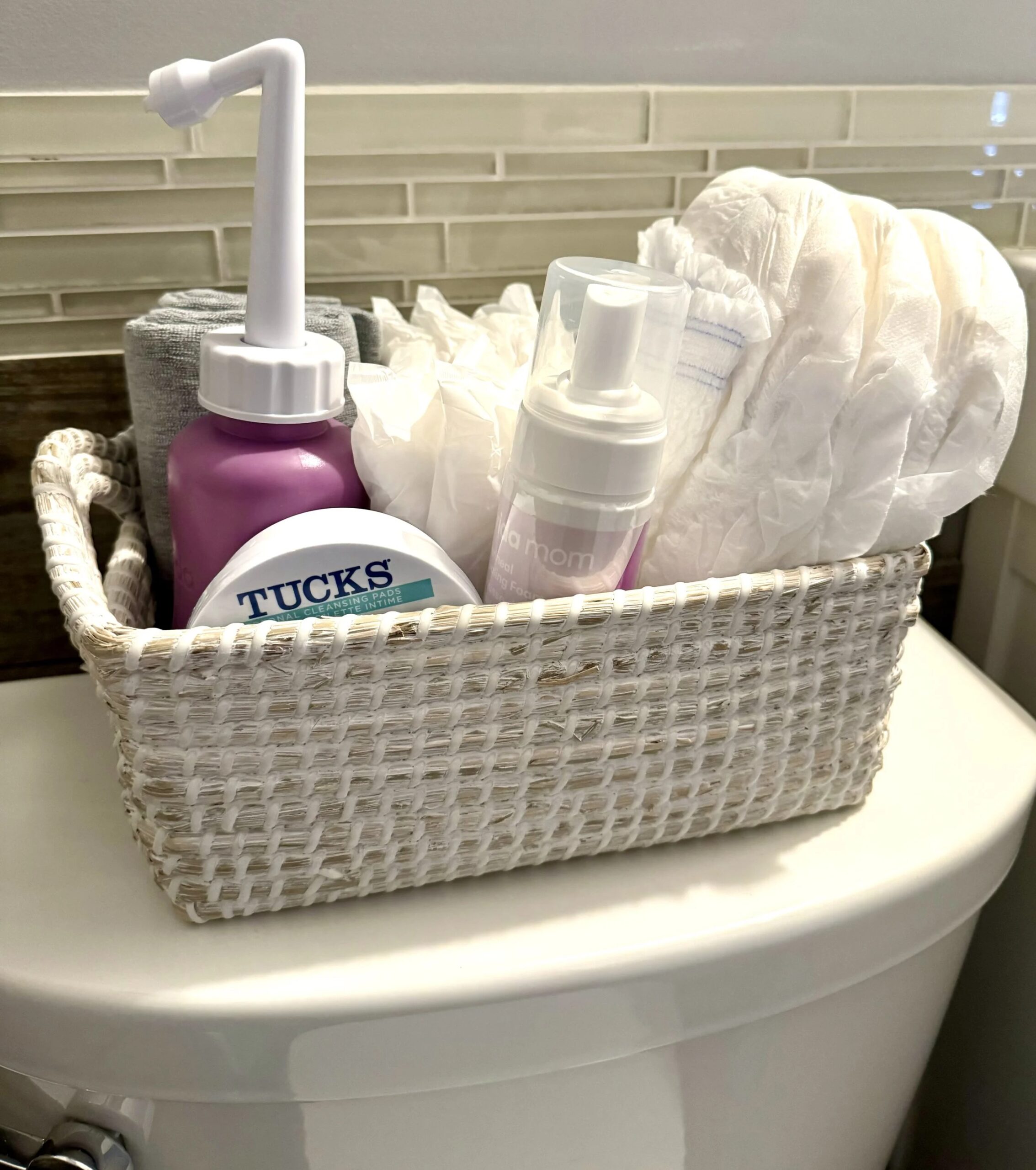 Basket of postpartum care supplies on a toilet tank, including a peri bottle, pads, wipes, and foam bottle
