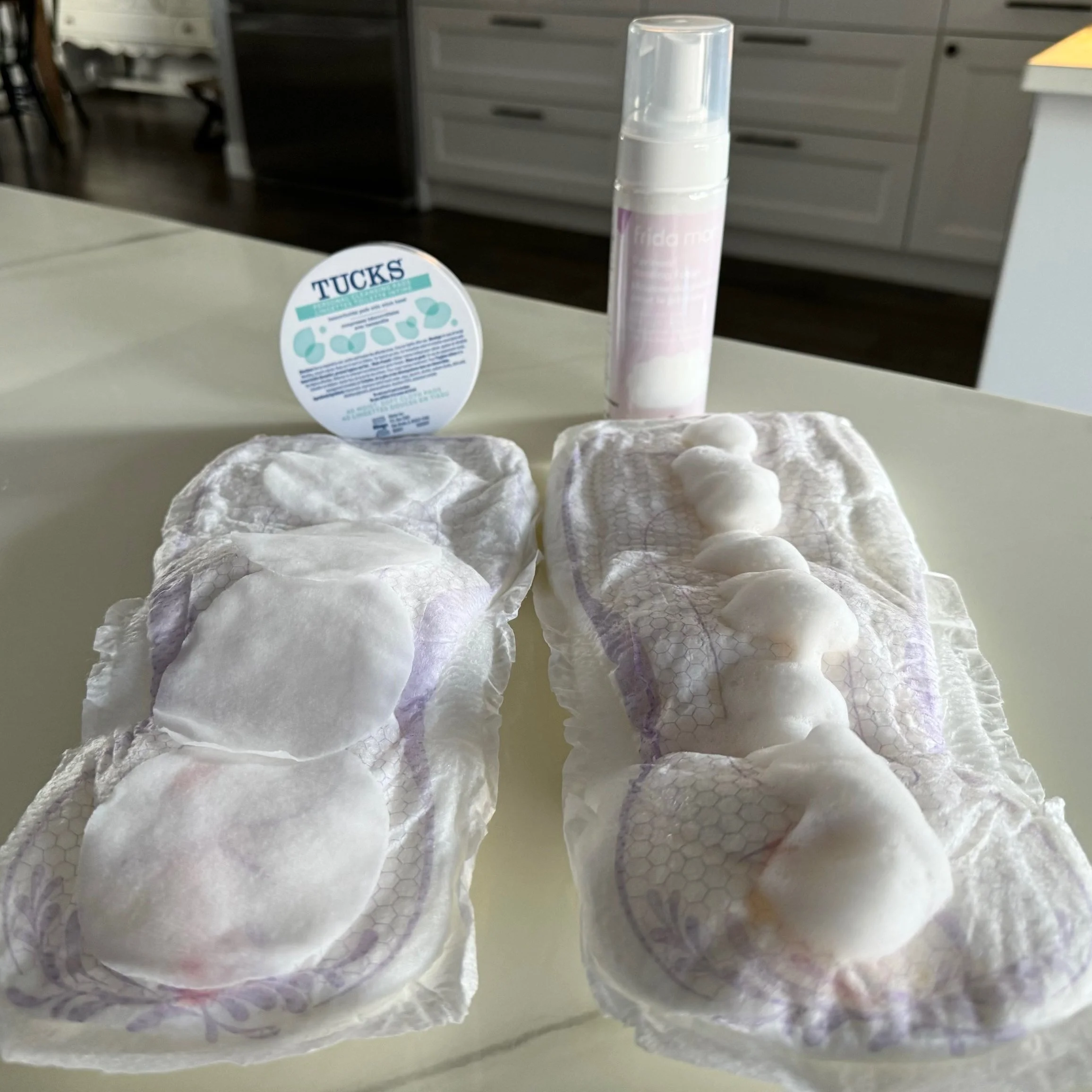 Two pads on a countertop with cotton rounds, foam, a Tucks container, and a pump bottle behind them