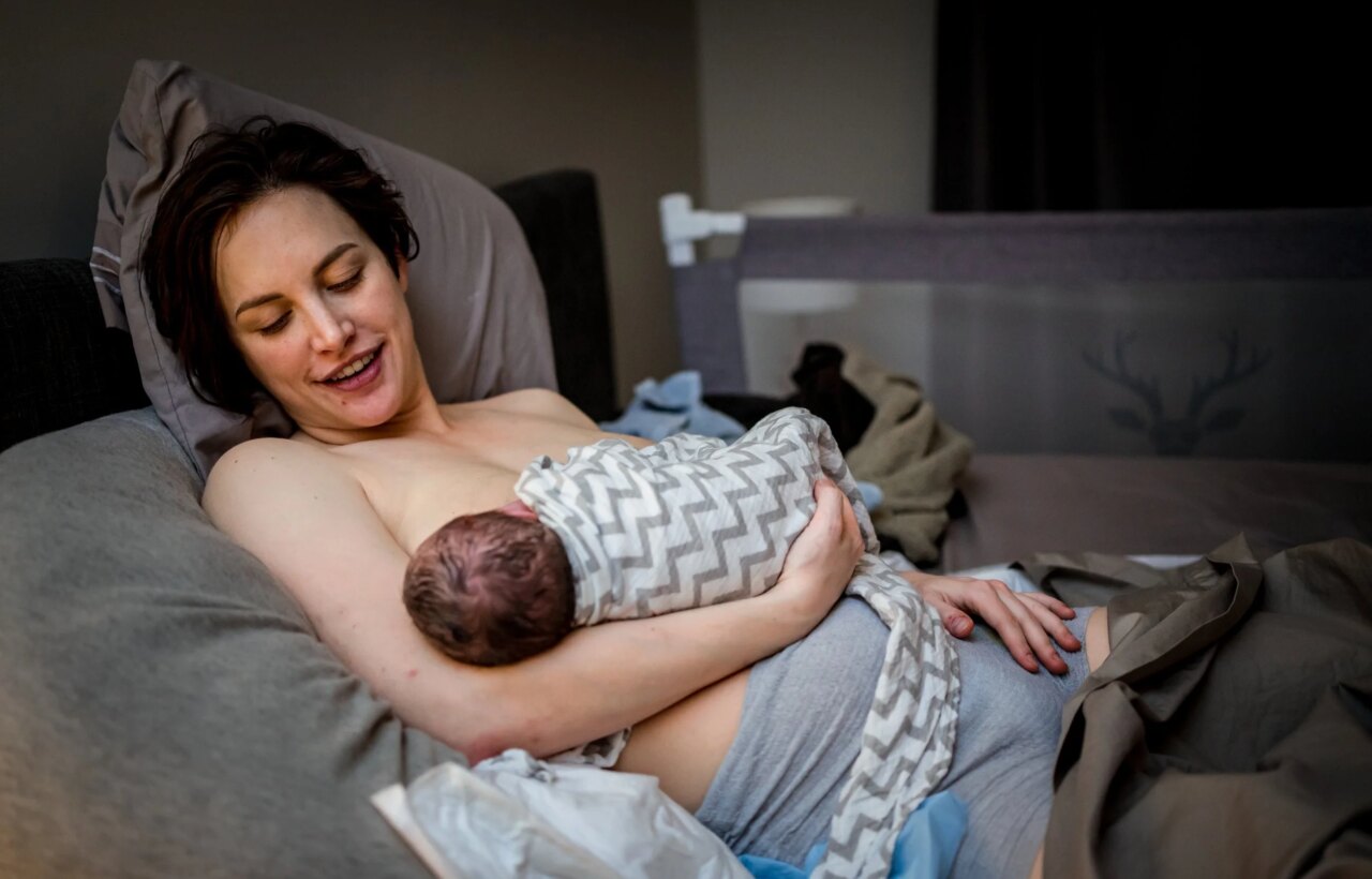 Adult reclines in bed holding a swaddled newborn close to their chest.