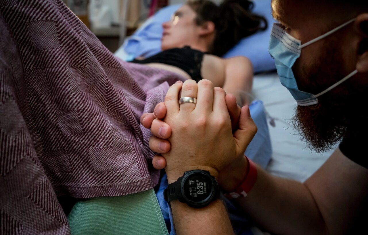 Masked person holds the hand of someone lying in bed under a patterned blanket.